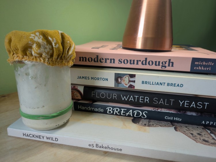 A jar of sourdough starter beside a pile of bakery cooking books including 'Modern Sourdough' by Michelle Eskeri, 'Brilliant Bread' by James Morton, and 'Flour Water Salt Yeast' by Ken Forkish.